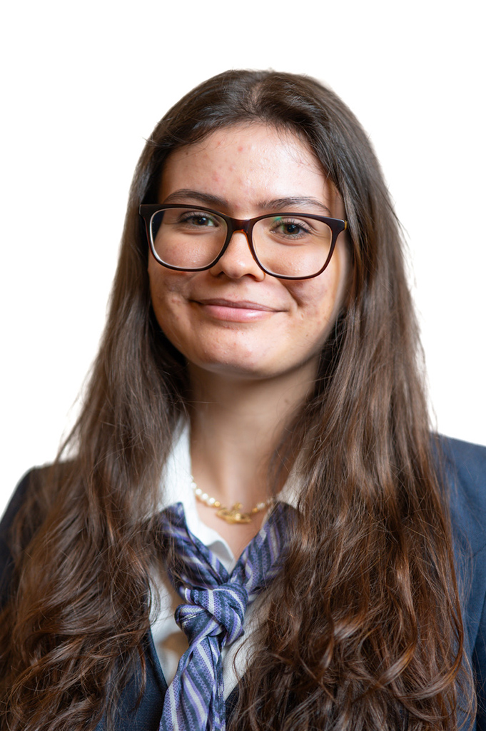 Eine junge Frau mit Brille lächelt, lange braune Haare, formelle Kleidung, neutraler Hintergrund.