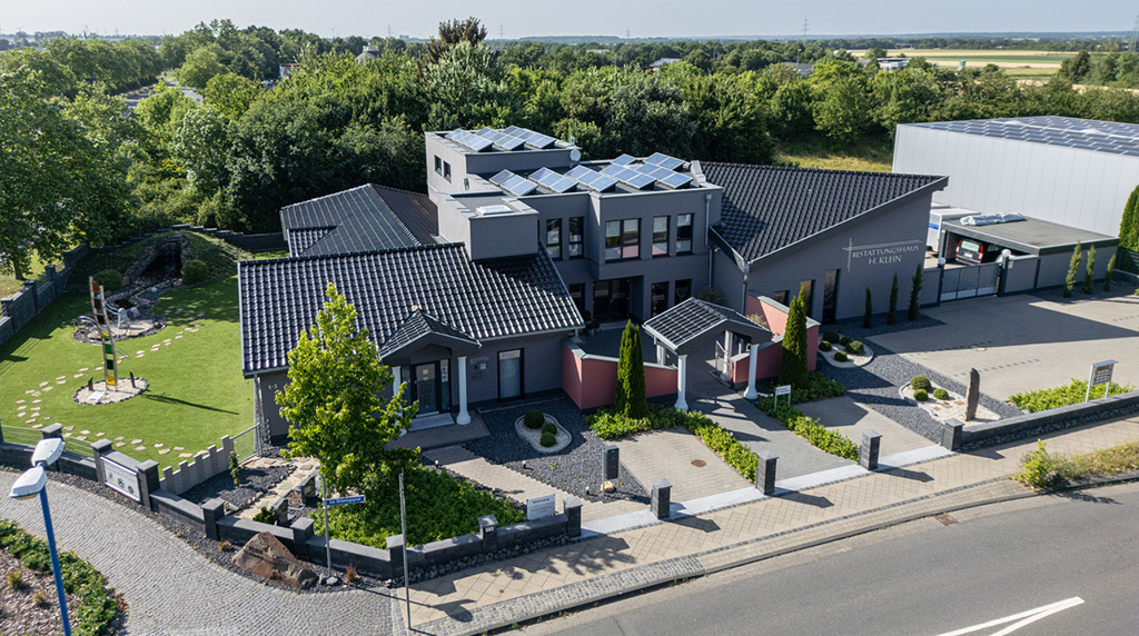 Moderne Gebäude mit dunkelgrauem Dach und Solarpanelen, umgeben von Garten und Bäumen.