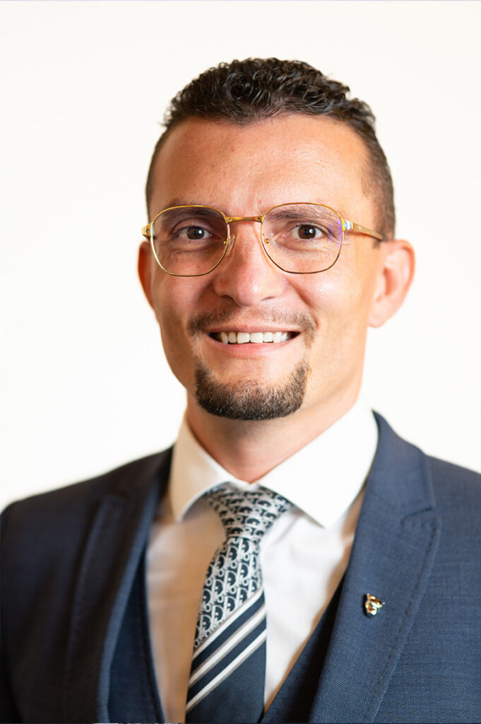 Ein Mann mit dunklem, kurzem Haar und Gläsern lächelt freundlich im Anzug